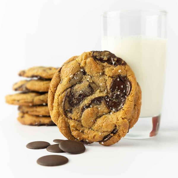 A New York Times chocolate chip cookie (David Leite), leaning against a glass of milk; a stack of cookies in back.