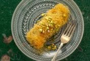 An rectangular dessert wrapped in thin stands of pastry, topped with crushed pistachios on a glass plate