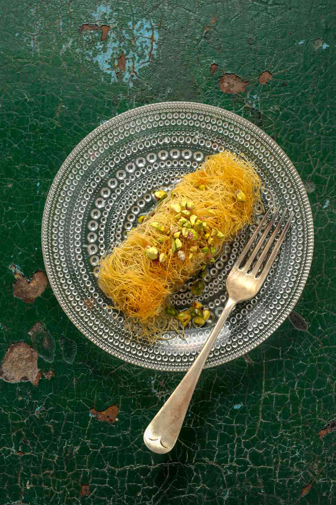 An rectangular dessert wrapped in thin stands of pastry, topped with crushed pistachios on a glass plate
