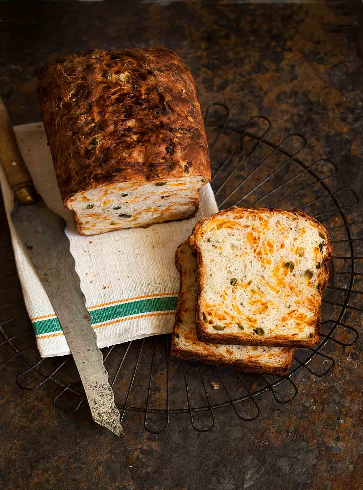 A loaf of no-knead Cheddar-chiles bread on a kitchen towel on a wire rack with two slices cut from it and knife resting beside it.