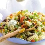 Confetti pasta salad in a large blue bowl with a wooden spoon, being drizzled with yogurt dressing