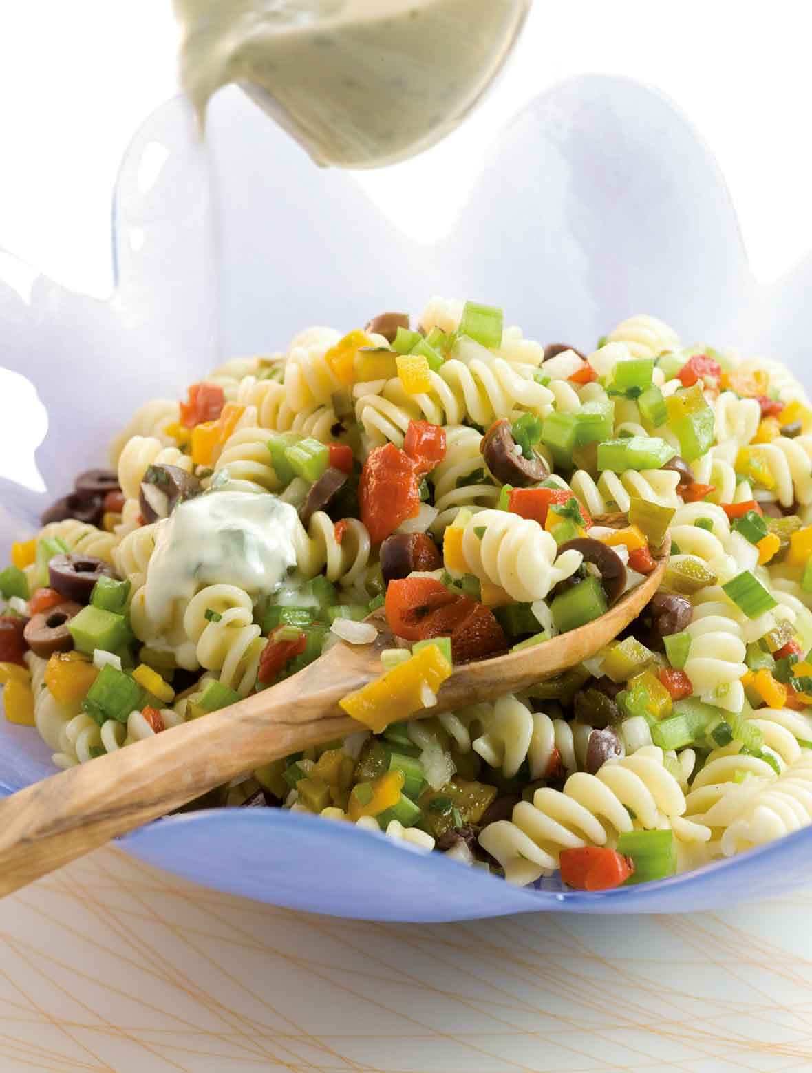 Confetti pasta salad in a large blue bowl with a wooden spoon, being drizzled with yogurt dressing