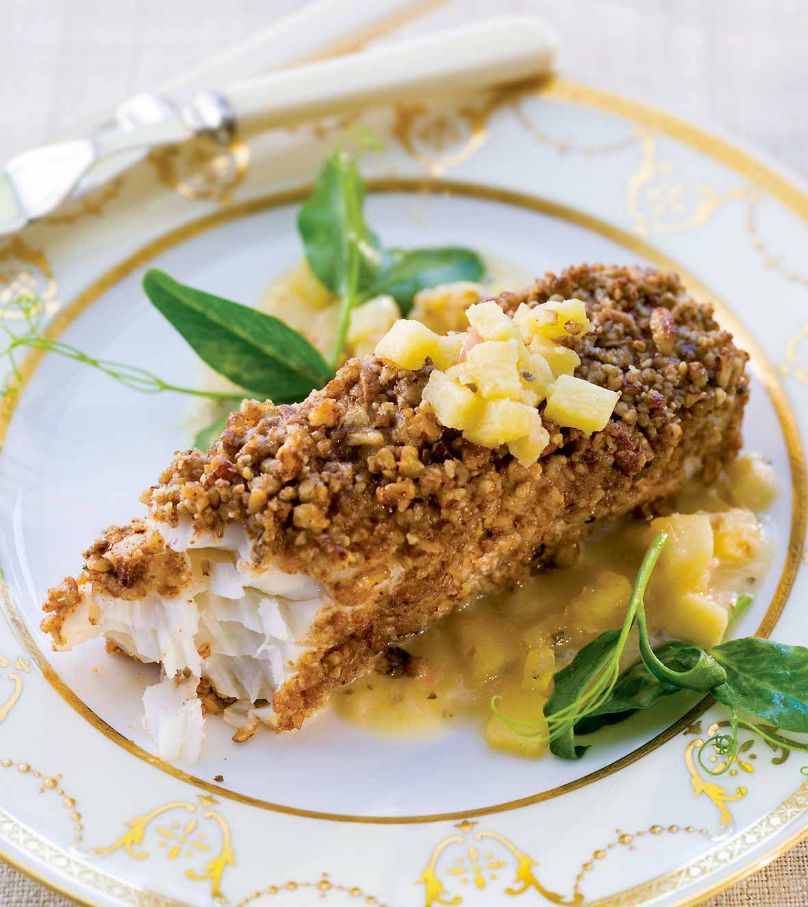 Halibut with nut crust, topped with apple vinaigrette, sitting on a plate with a knife and fork.