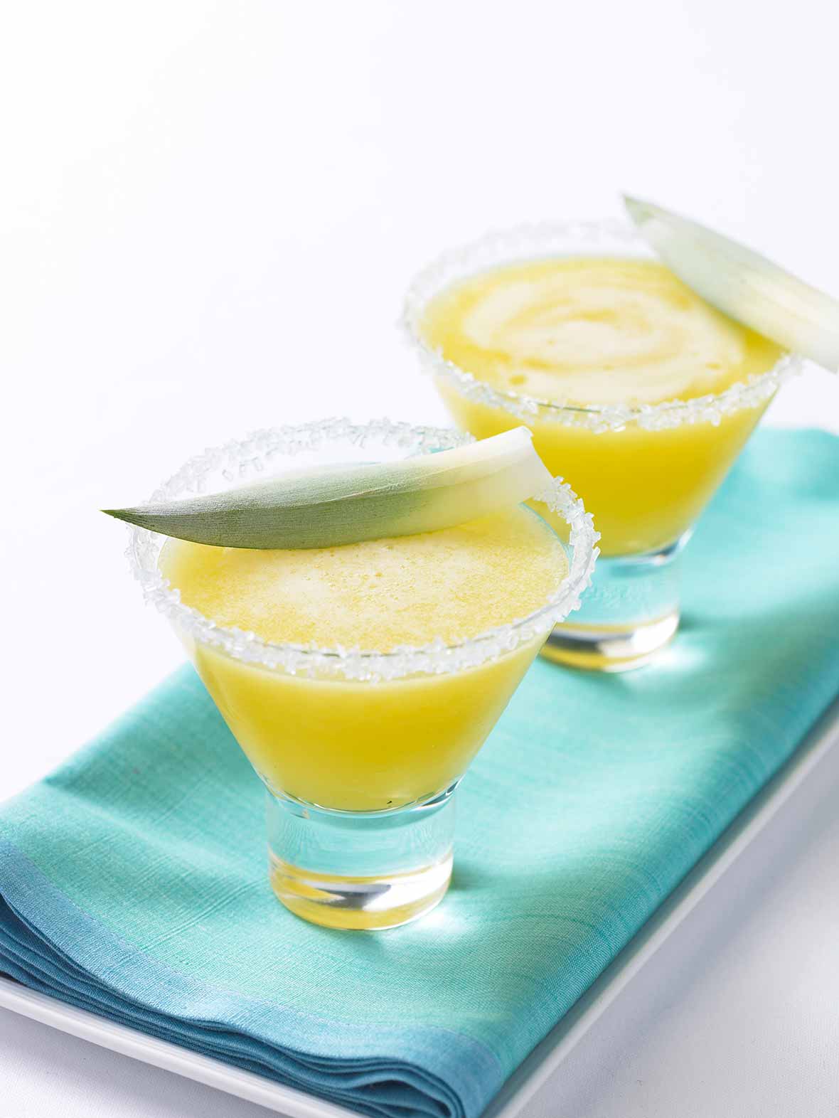 Two glasses filled with pineapple-tequila smoothie, rimmed with sugar, topped with line wedges on a napkin