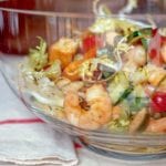 A glass bowl filled with cooked shrimp, grilled focaccia, cucumbers, tomatoes, capers, olives, and frisee.
