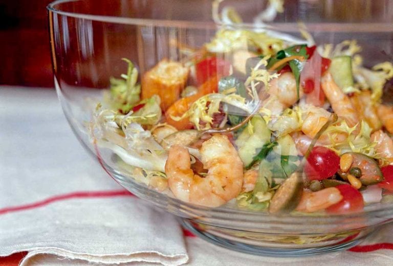 A glass bowl filled with cooked shrimp, grilled focaccia, cucumbers, tomatoes, capers, olives, and frisee