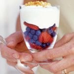 Zabaglione with summer fruit layered in a glass, topped with crushed biscotti, being held by two hands.