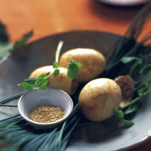 Three Chinese tea eggs in a black bowl with chives, pea shoots, and a small bowl of sesame seeds.