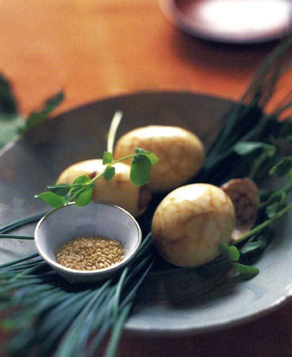 Three Chinese tea eggs in a black bowl with chives, pea shoots, and a small bowl of sesame seeds.