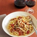 A wide white bowl filled with fettuccine with a savory veal sauce and a carafe and glass of wine in the background.