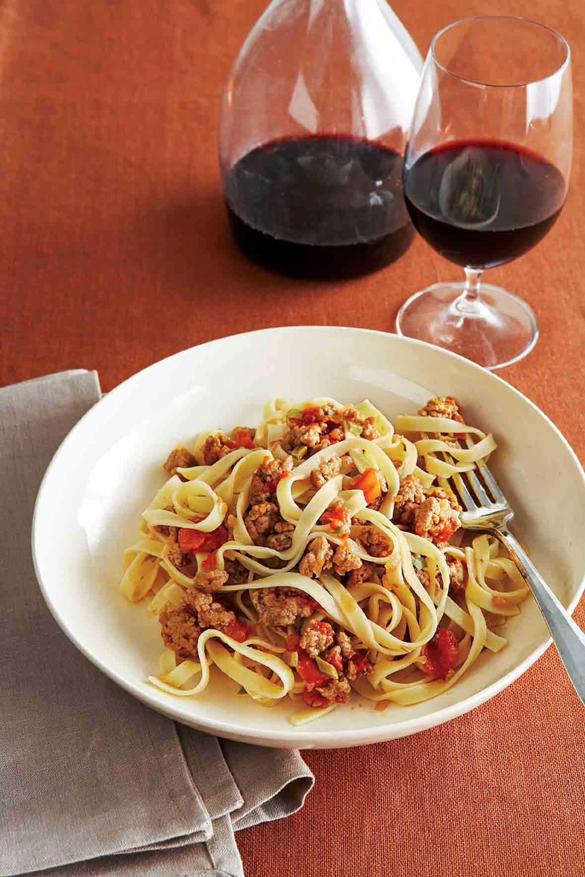 A wide white bowl filled with fettuccine with a savory veal sauce and a carafe and glass of wine in the background.