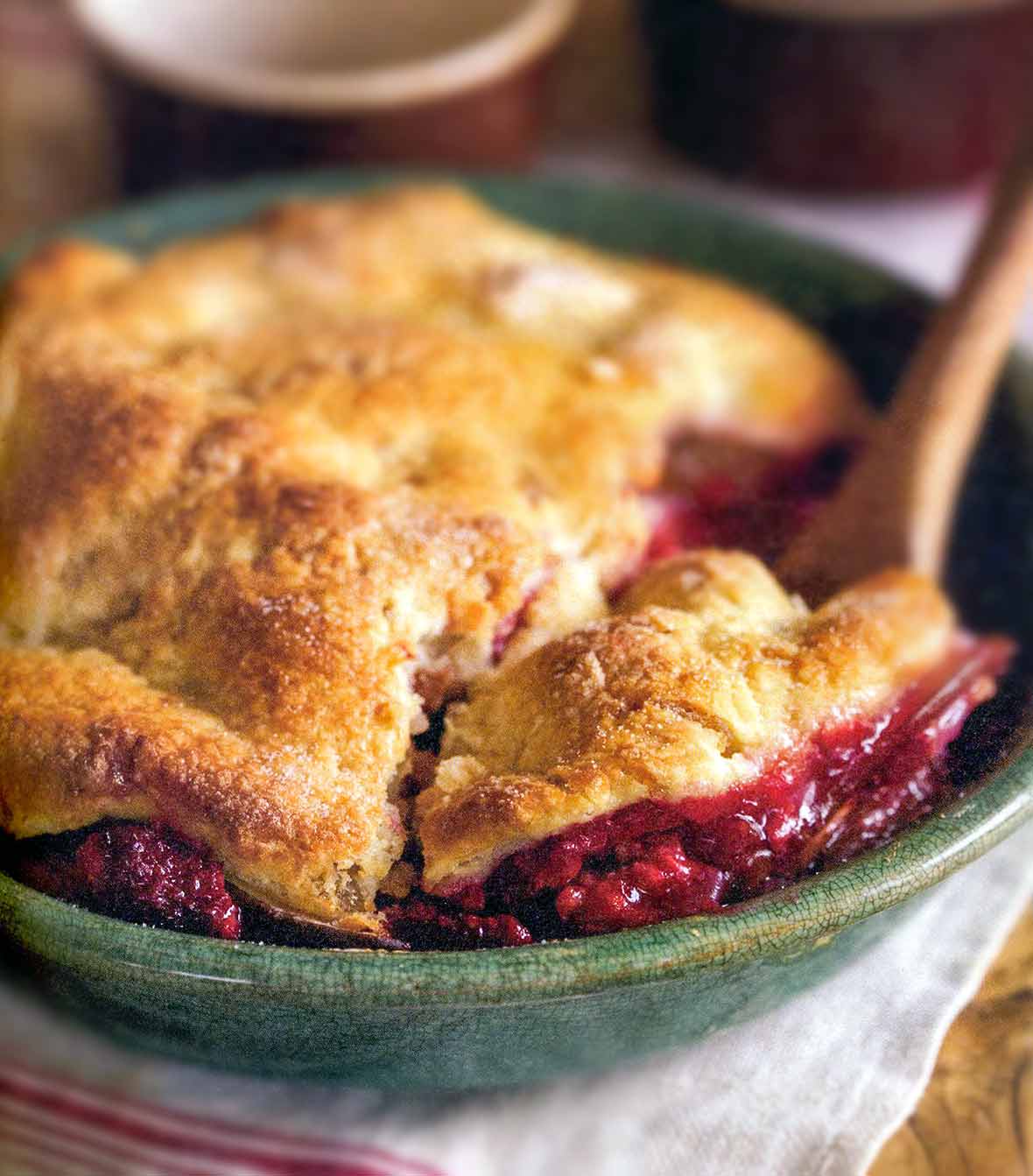 A pear and raspberry pandowdy or cobbler in a green bowl with a pastry top, wooden spoon inside.