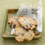 Irregular-shaped wafer cookies dotted with lemon and black olives in a wax paper wrapper on a green wood tray.