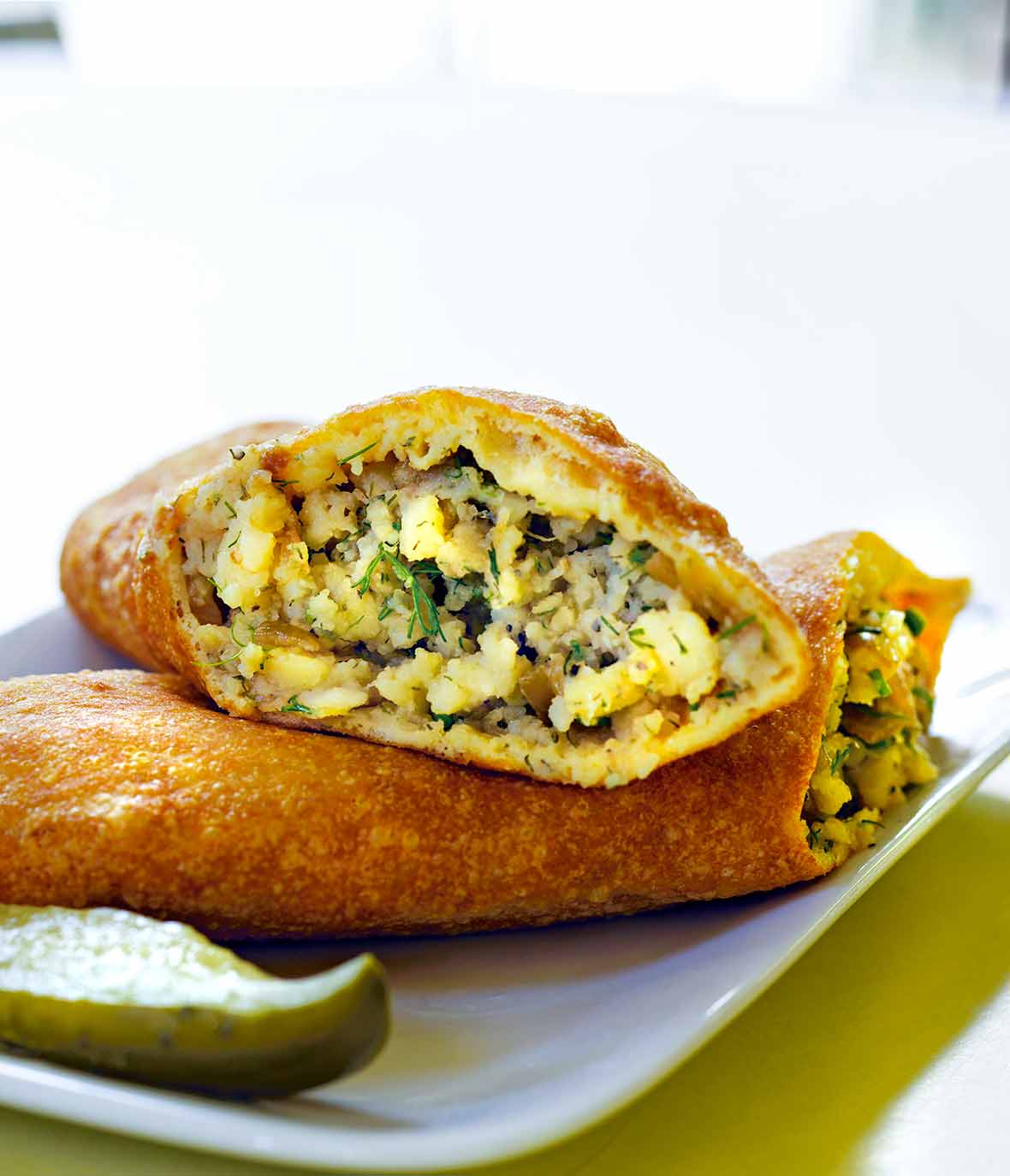 A potato piroshki cut in half, revealing the filling, on a plate with a pickle wedge