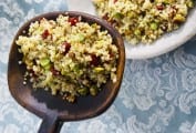 A large wooden spoon with a scoop of quinoa salad with pistachios and cranberries being held over a white bowl filled with the salad.