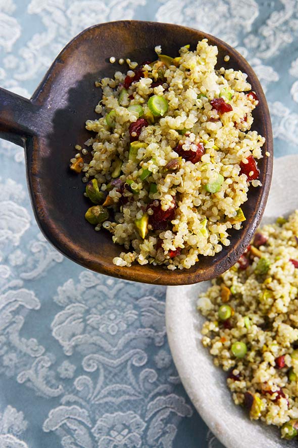 A large wooden spoon with a scoop of quinoa salad with pistachios and cranberries being held over a white bowl filled with the salad.