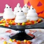Four meringue ghosts on a bed of candy corn on a black cake stand.