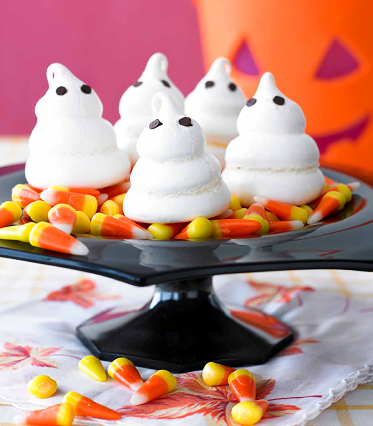 Four meringue ghosts on a bed of candy corn on a black cake stand.