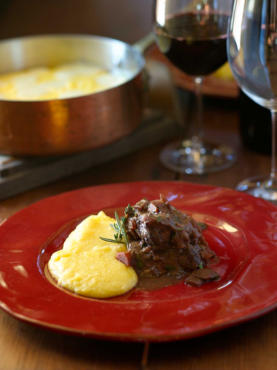 Rabbit in red wine, Ischia style, on a red plate with a glass of red wine and a pan of polenta in the background