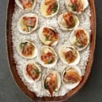 Clams casino, covered with slices of bacon and herbed butter, sit in a large pottery dish filled with rock salt.