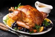 An old-fashioned roast turkey on a platter with bunches of grapes and orange slices and a boat of gravy behind it.