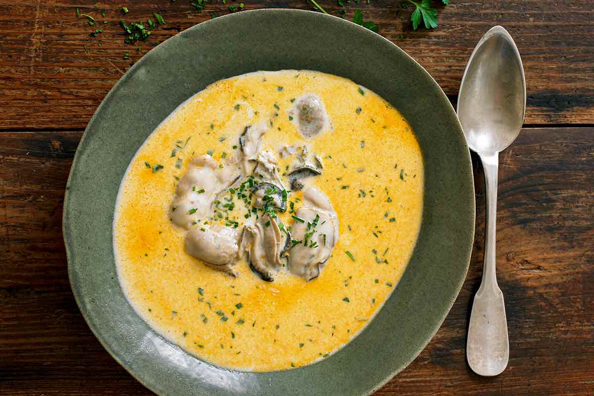 A ceramic bowl filled with oyster stew with several oysters in the center and a spoon resting beside the bowl.