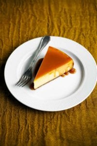 A slice of pure pumpkin cheesecake on a white plate drizzled with caramel glaze with a fork resting beside it.