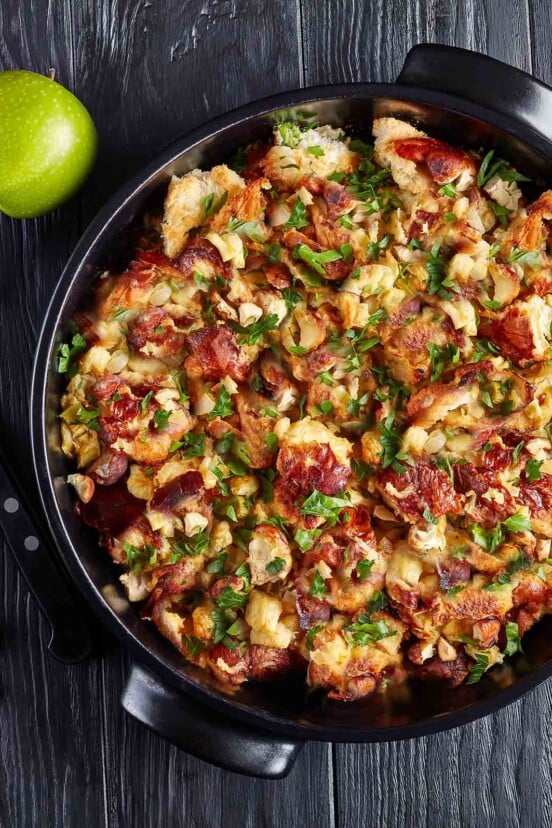 A black bowl of bread stuffing with herbs and bacon, alongside is a green apple.