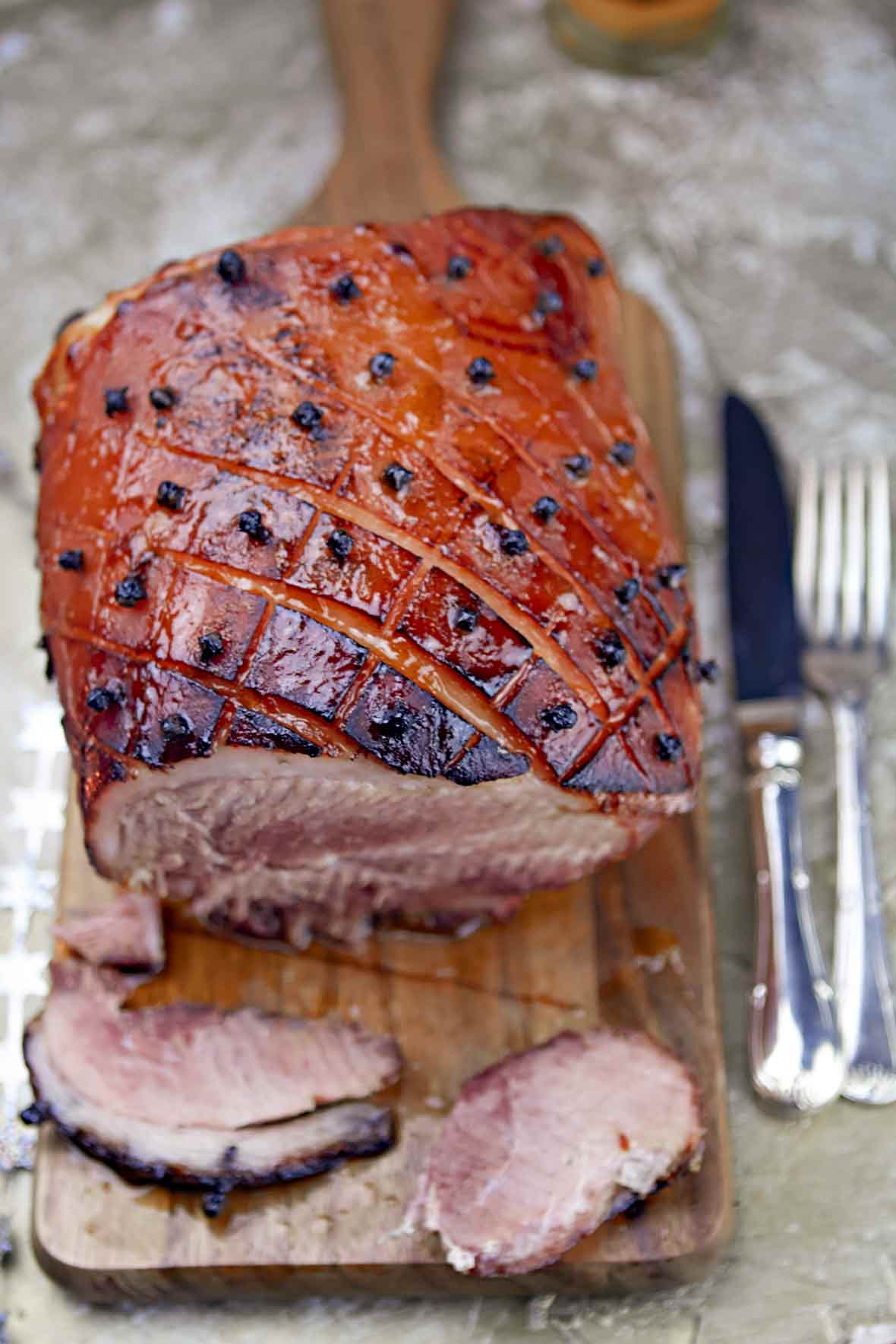 A root beer-glazed ham on a wooden cutting board with a fork and knife resting beside it.