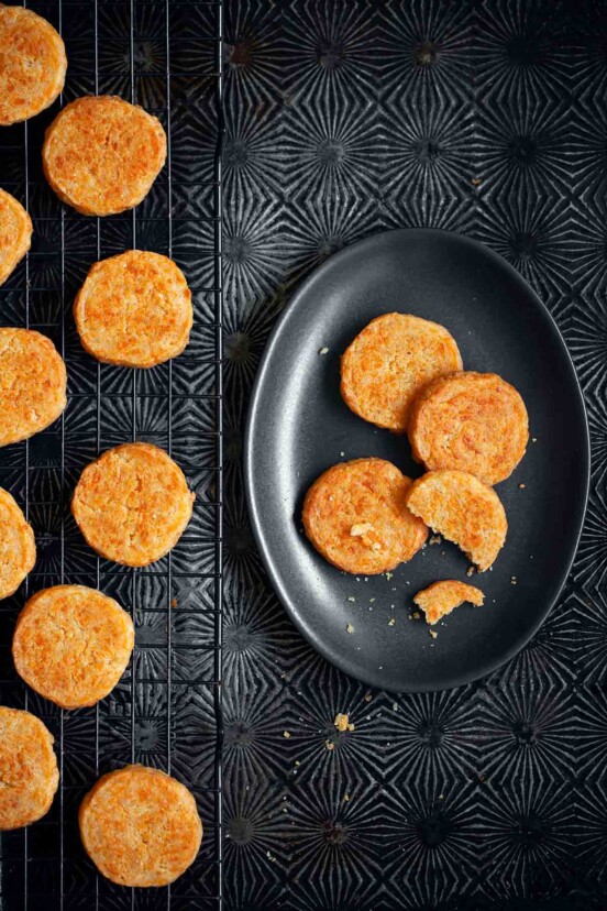Cheddar-Parmesan crackers on a black wire rack with a few on a black oval plate beside the rack.