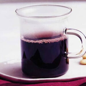 A glass mug of glogg aquavit on a white saucer with a pile of almonds on a red placemat.