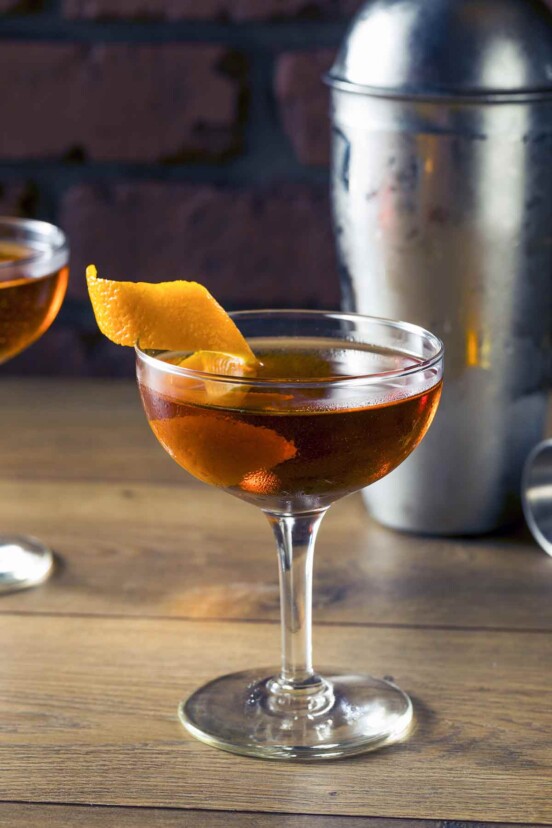 Two coupe glasses filled with martinez cocktail and a strip of orange zest with a cocktail shaker and more orange zest in the background.