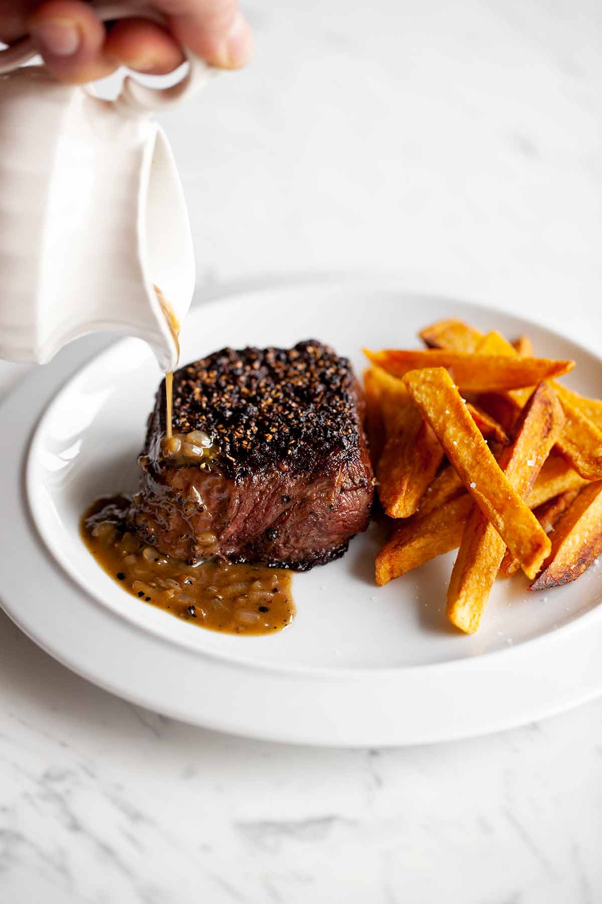 A person drizzling sauce over a steak au poivre on a white plate with sweet potato fries.