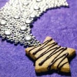 A walnut star, drizzled with dark chocolate ganache, on a purple background next to a sparkly moon ornament.
