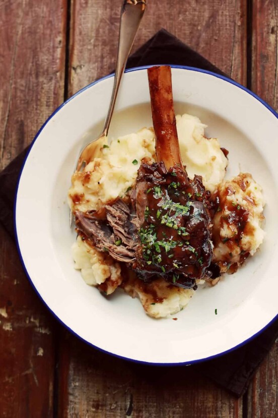A braised lamb shank in pinot noir, and exposed bone, on mash potatoes, all on a white plate sitting on planks of wood