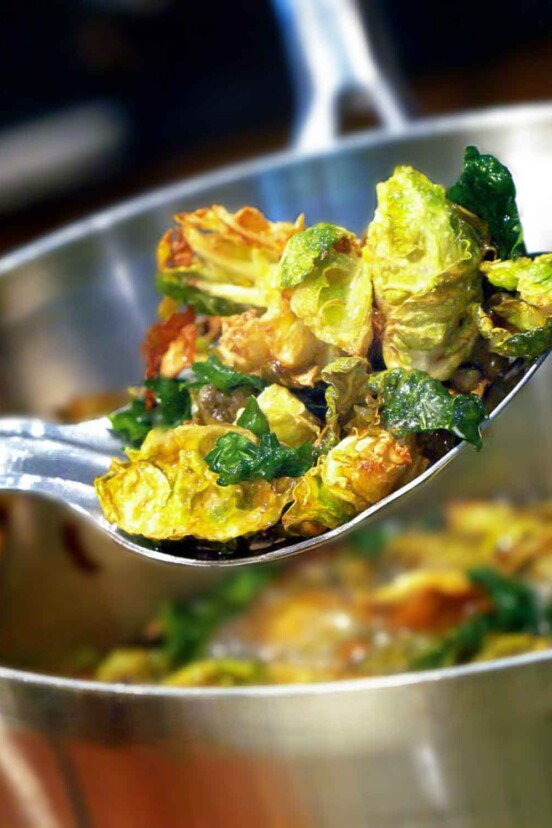 A serving spoon of fried Brussels sprouts with walnuts and capers resting over a large metal bowl.