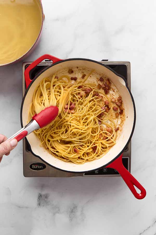 Cooked spaghetti being added to pancetta in skillet for carbonara.