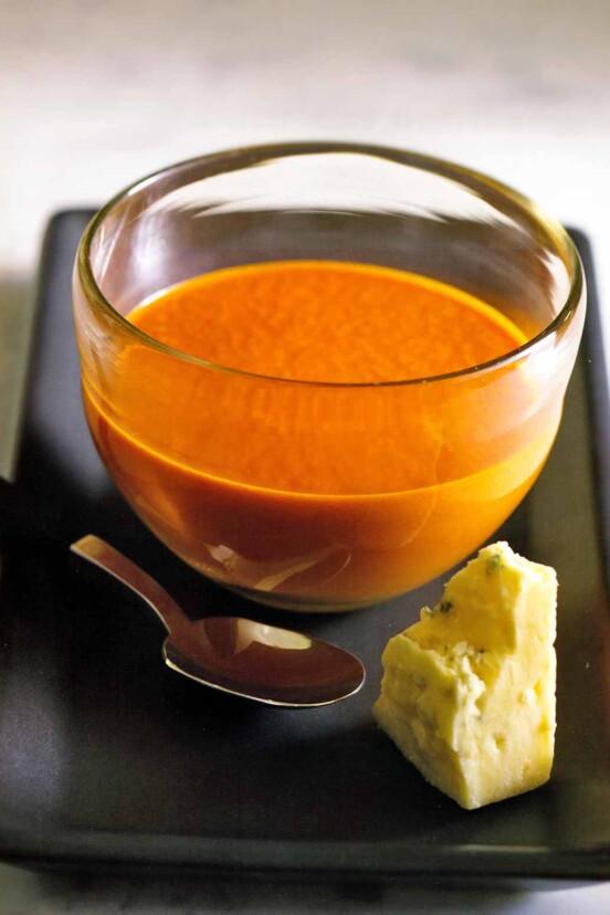 Glass bowl with spicy tomato blue cheese soup on a tray with a hunk of cheese and a spoon.
