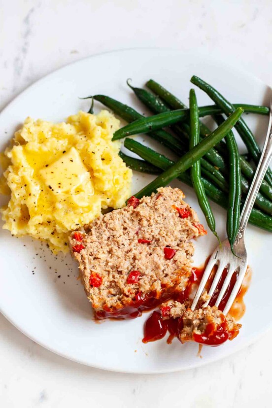 A slice of turkey meatloaf on a white plate with mashed potatoes, green beans, and fork resting on the side.