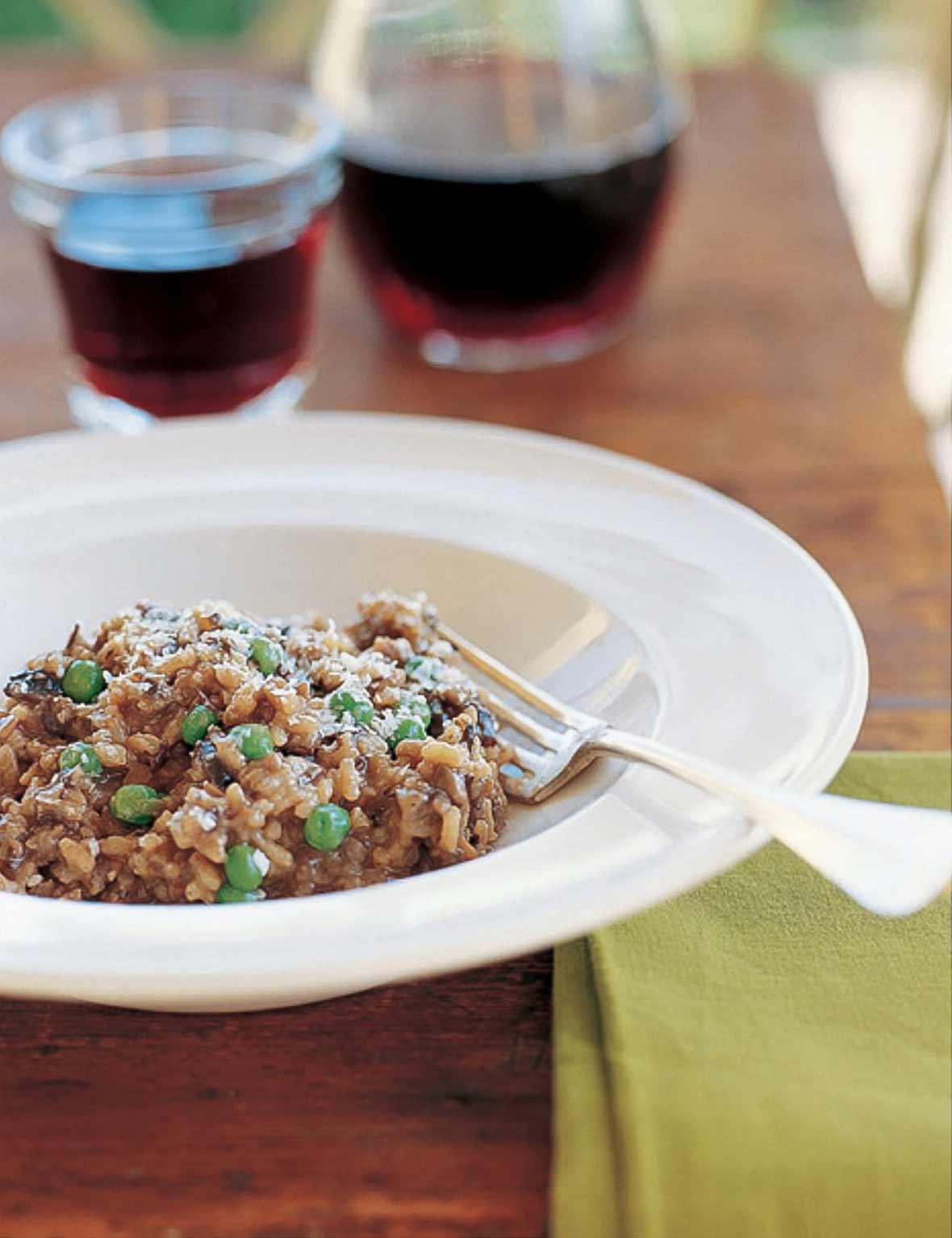 A white bowl of wild mushroom risotto with pea with a fork resting inside and a glass and decanter of wine in the background.