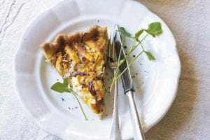 A slice of butternut squash and Parmesan tart on a white plate with a fork and knife, garnished with microgreens.