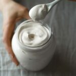 A person holding a spoonful of milk mayonnaise above a jar.