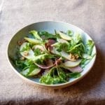An arugula salad with country ham, pears, and honey vinaigrette, topped with chopped walnuts in a white bowl.