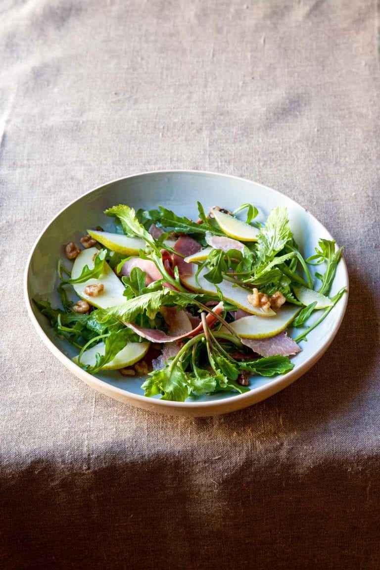 An arugula salad with country ham, pears, and honey vinaigrette, topped with chopped walnuts in a white bowl.