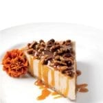 A slice of banana rum cheesecake with maple rum sauce and pecan pieces on top and a marigold beside the cake.