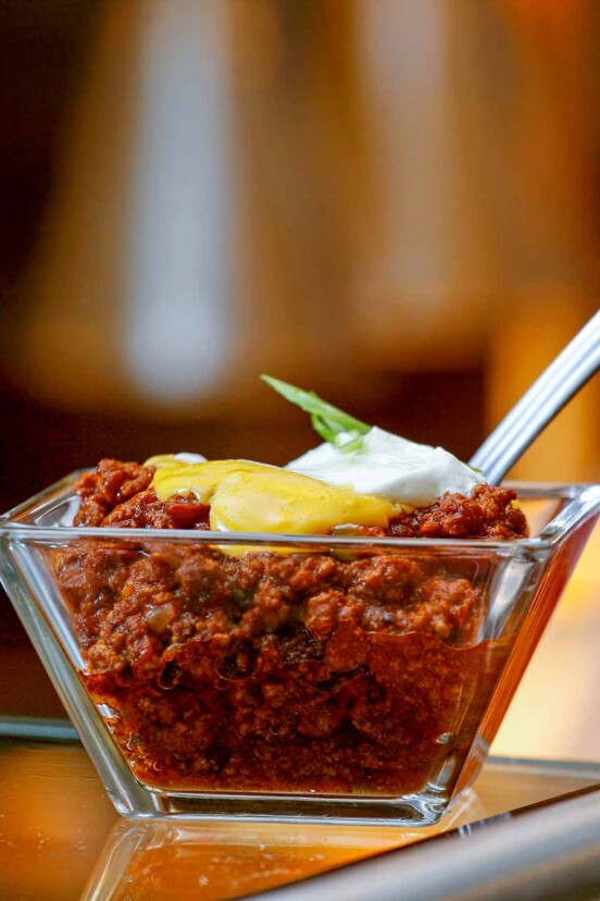 A glass dish of beef chili topped with cheddar cheese sauce and a dollop of sour cream with a spoon resting inside.
