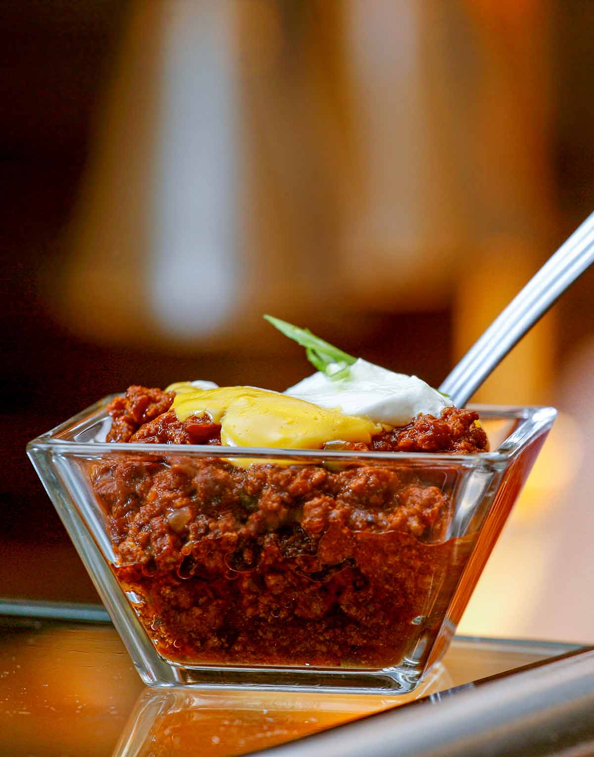 A glass dish of beef chili topped with cheddar cheese sauce and a dollop of sour cream with a spoon resting inside.