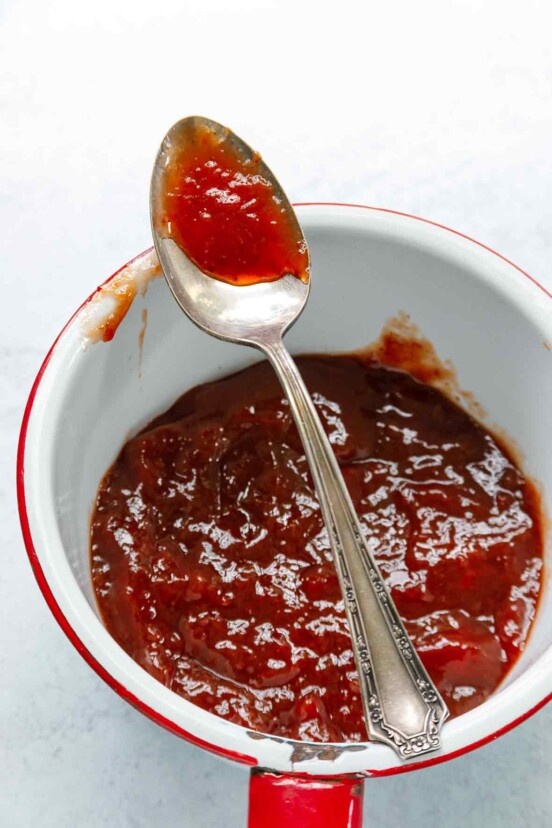 An enamel-coated pot filled with strawberry jam and a spoon resting on top.