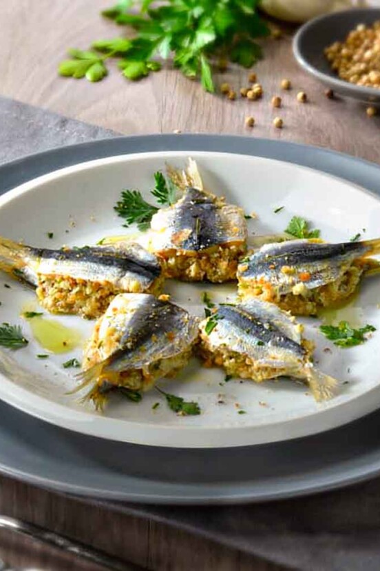 Five stuffed sardines on a dinner plate with pine nuts and parsley in the background