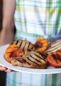 A woman holding a white platter of grilled country pork chops with grilled peaches.